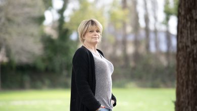 A woman with short blonde hair stands in a park outdoors. She is wearing a striped t-shirt, jeans, a black cardigan and a silver cross around her neck
