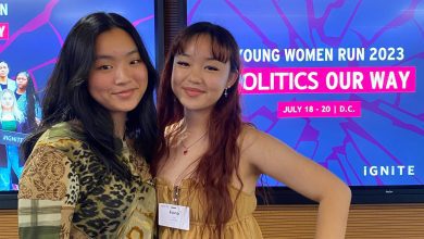 A photo of two teenagers posing for a photo together. They stand in front of a screen that reads, "Women run 2023. Politics our way."